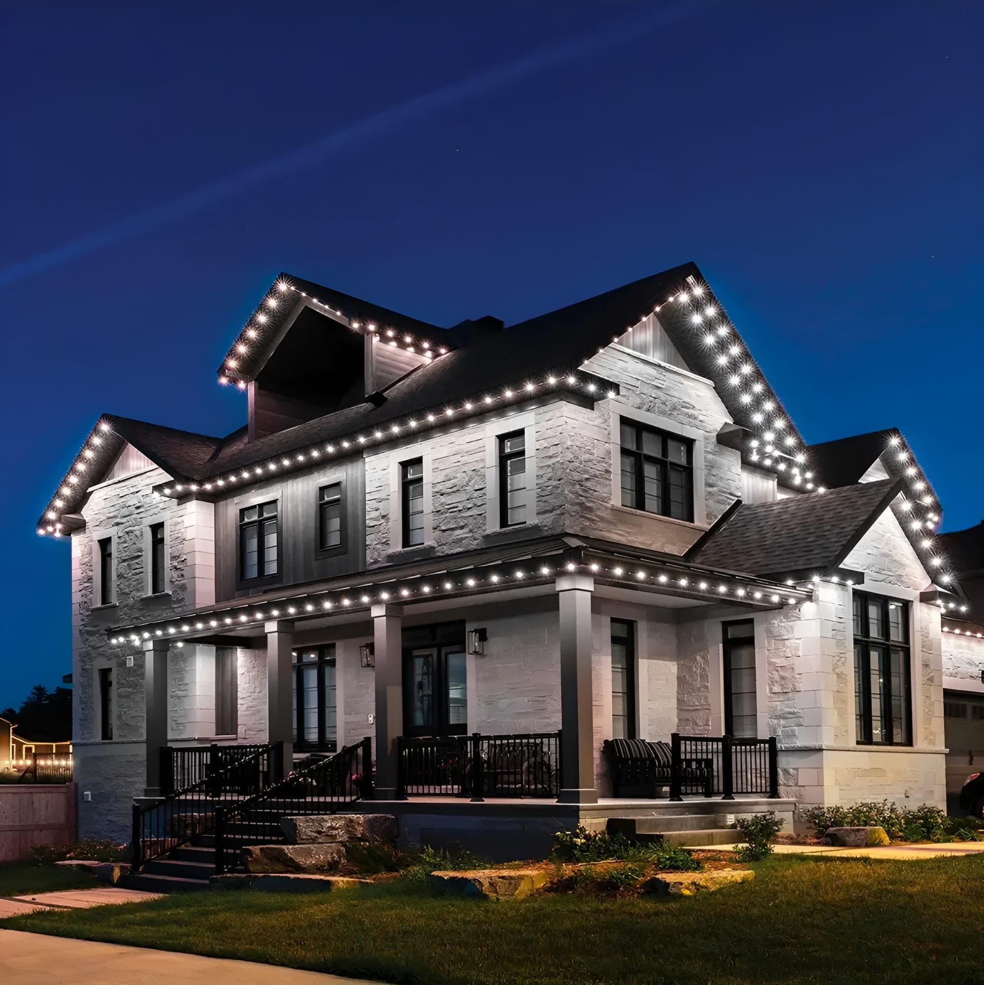 Cool white permanent roofline lights outlining the peaks, dormers and wraparound porch of a stone two-storey home at night