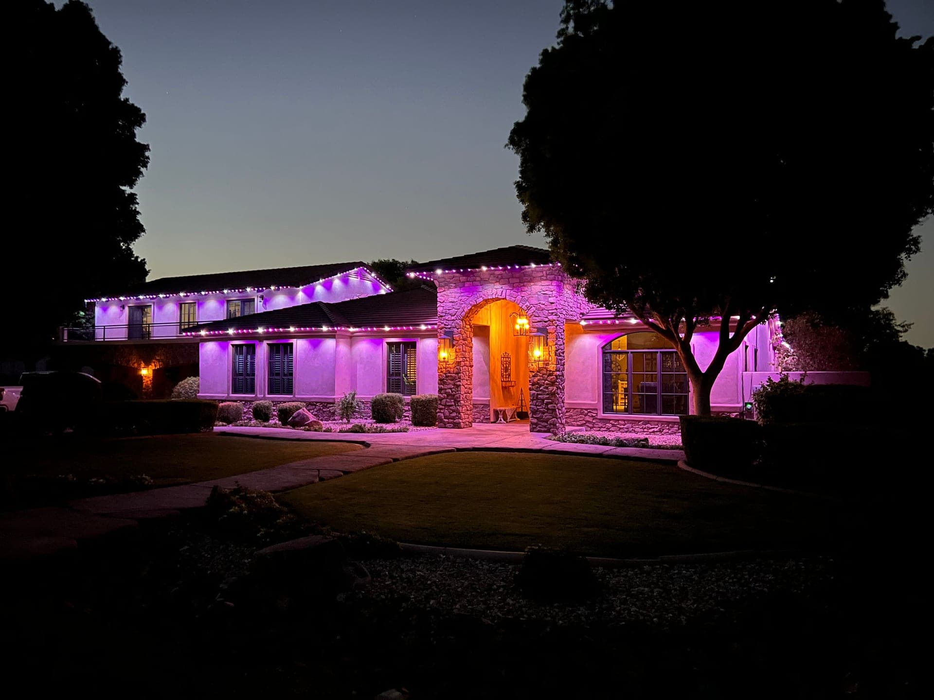 Pink and purple permanent roofline lights on a stucco ranch home with a lit arched entry at dusk