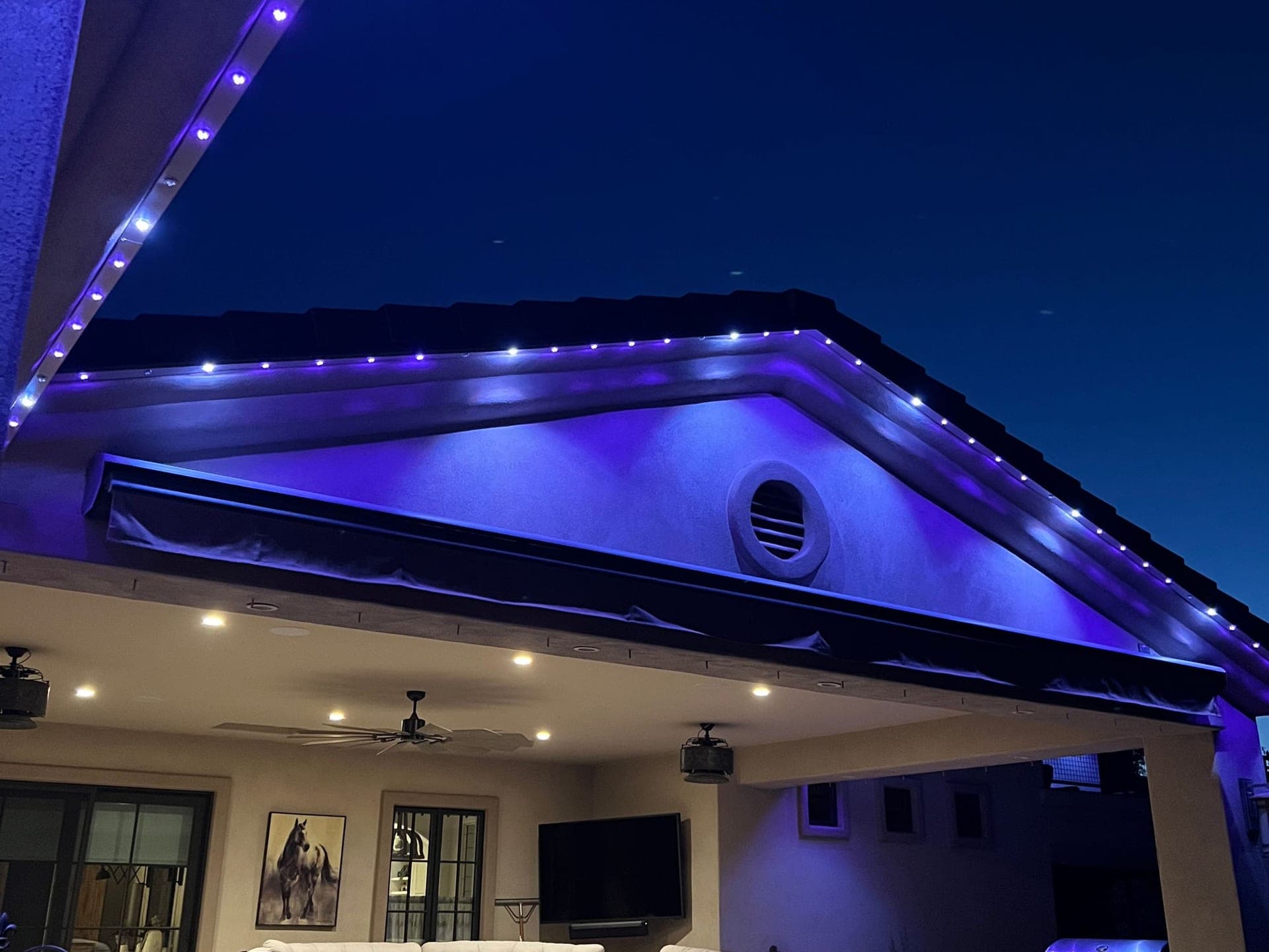 Purple permanent roofline lights along the gable of a stucco home above a covered patio, photographed from below