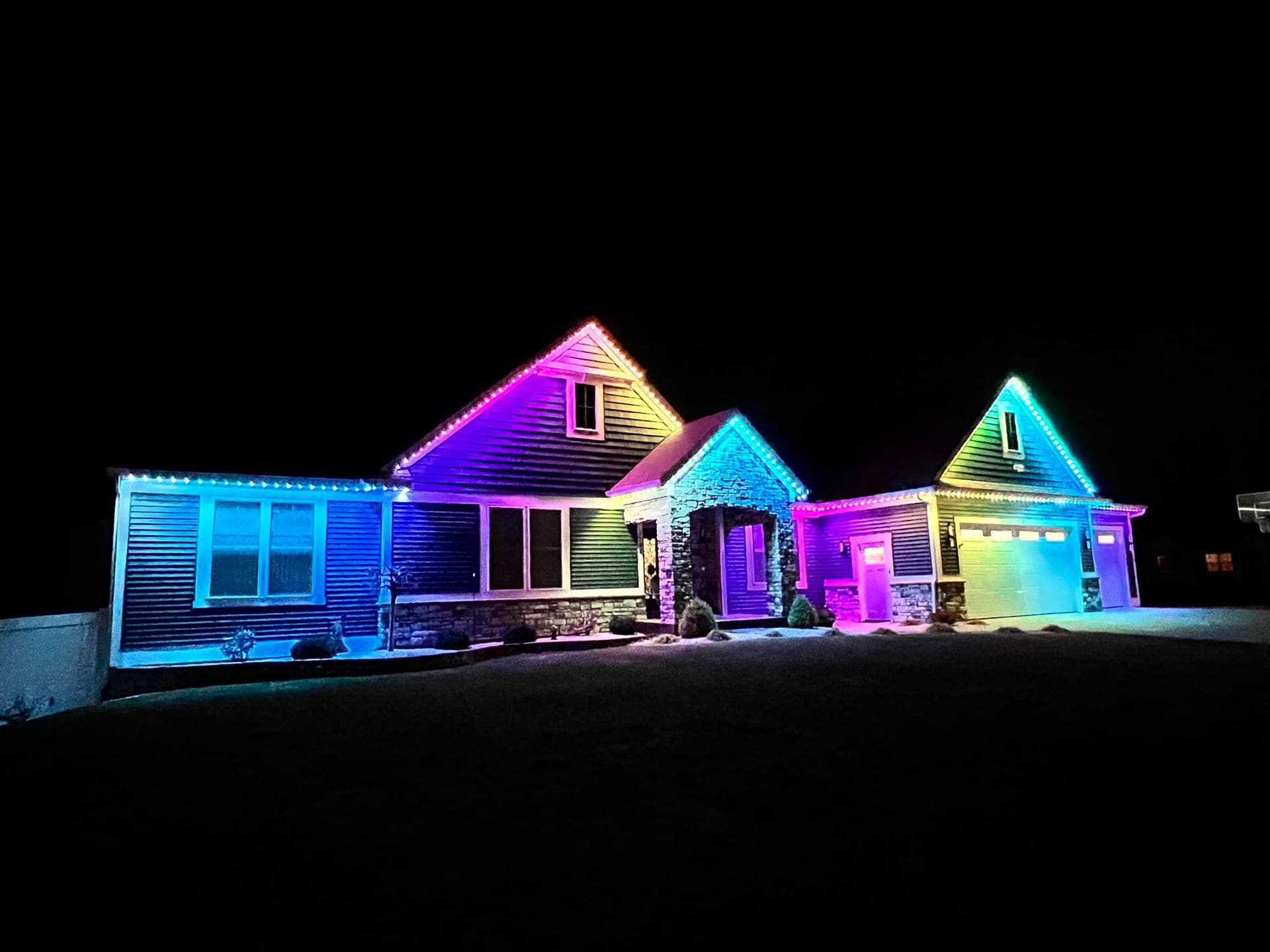 Rainbow gradient permanent roofline lights across the triple gables of a dark-sided bungalow with stone accents