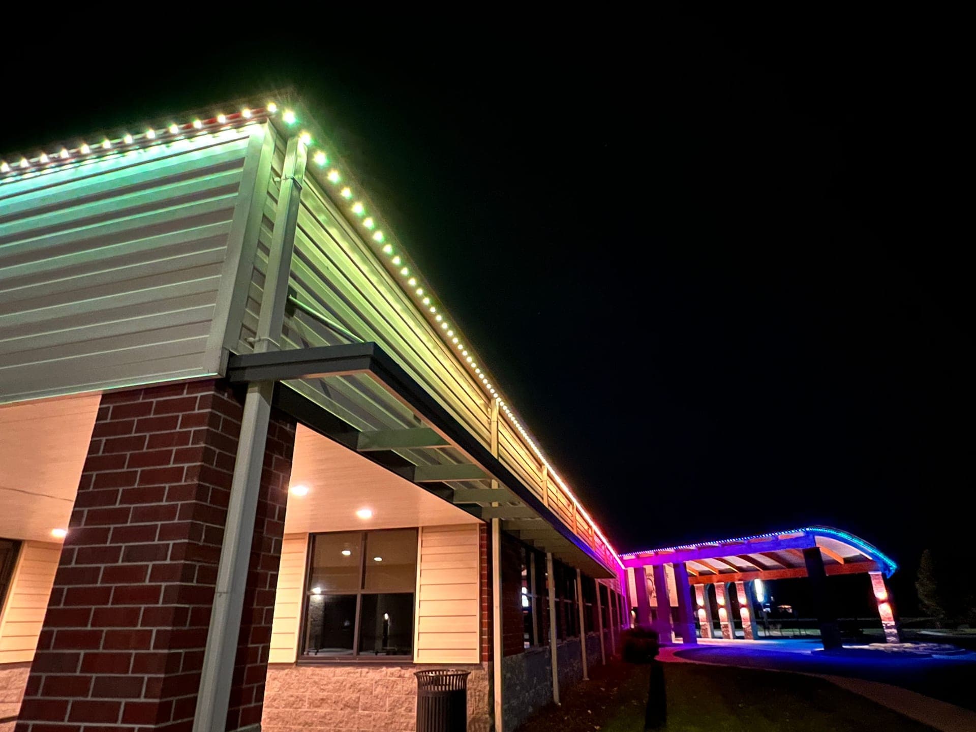 Rainbow permanent roofline lighting along the eaves of a brick commercial building with a covered entry canopy