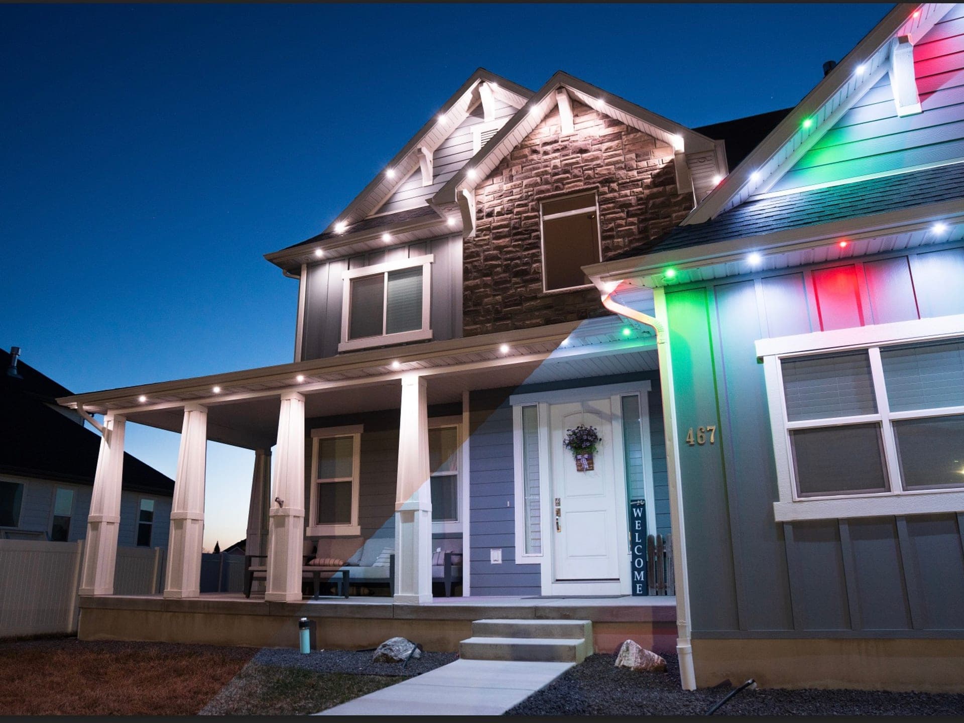 Red, white and green permanent Christmas roofline lights on a blue two-storey home with a stone gable and white porch