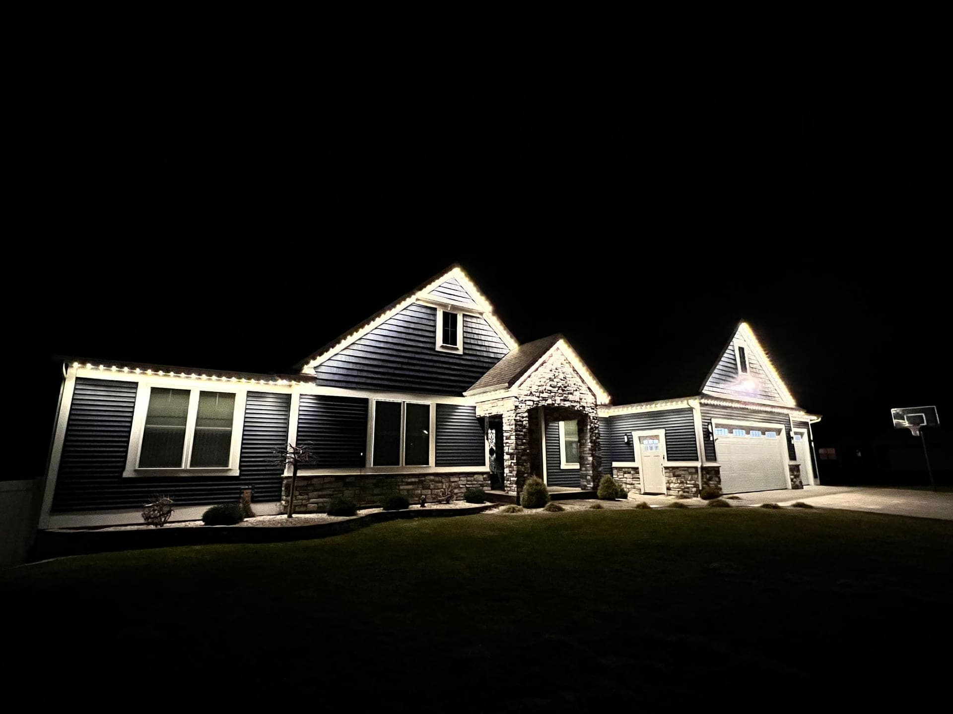 Warm white permanent roofline lights on a dark-sided bungalow with three front gables and a stone entry