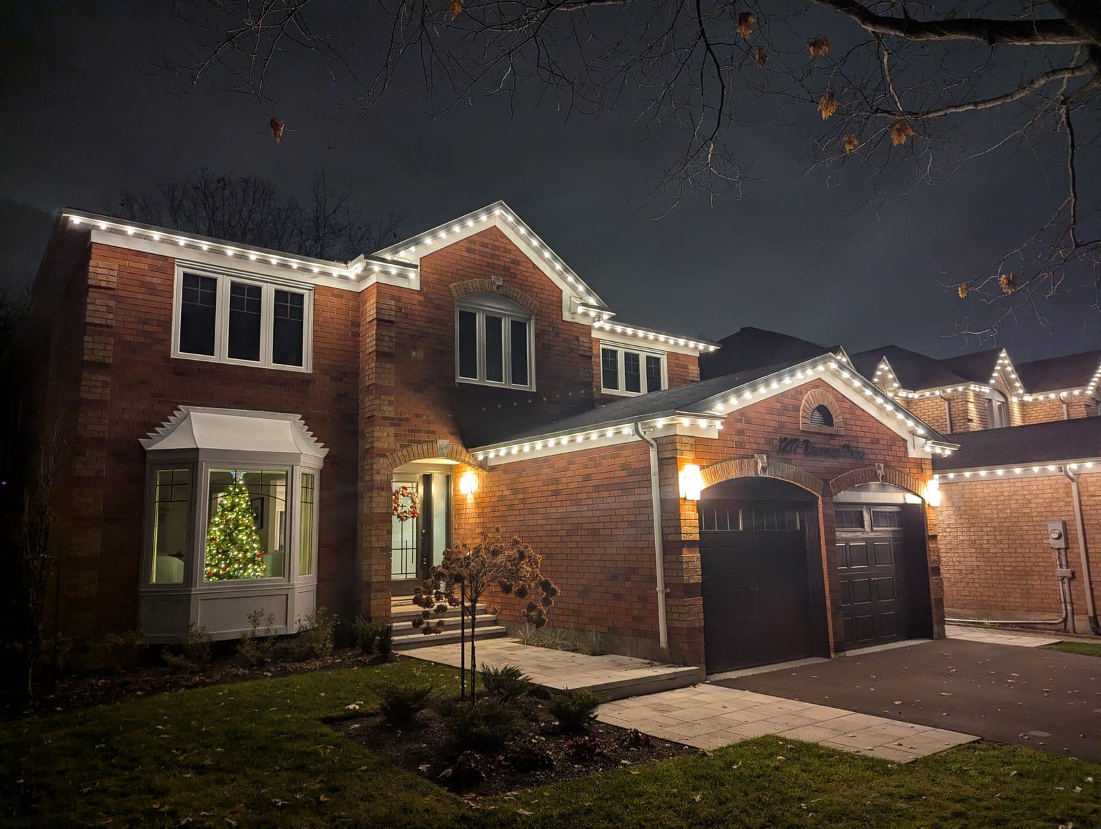 Brick two-storey home with warm-white roofline lights and a Christmas tree glowing in the bay window