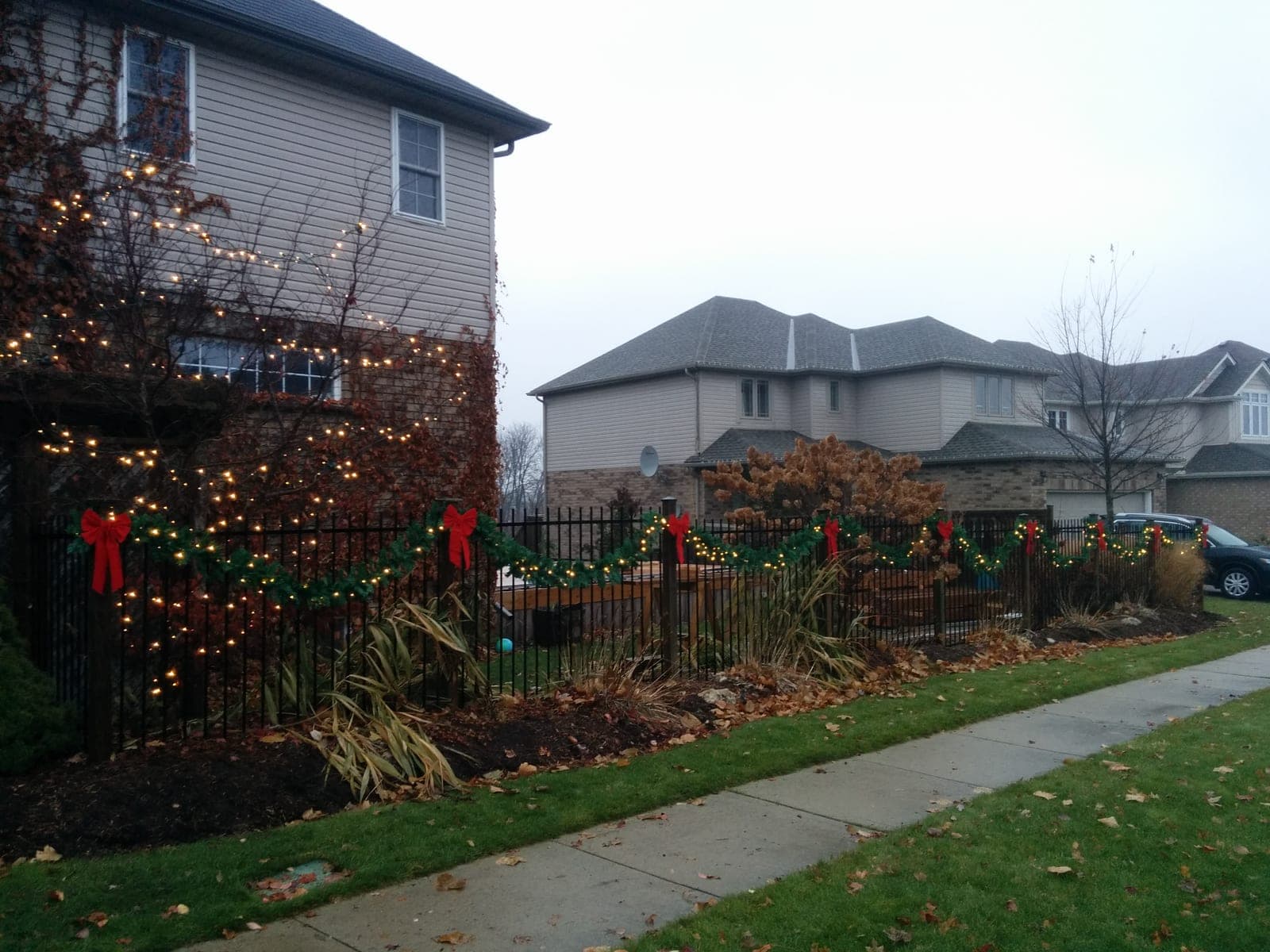 Front-yard iron fence draped in lit Christmas garland with red bows beside a decorated home