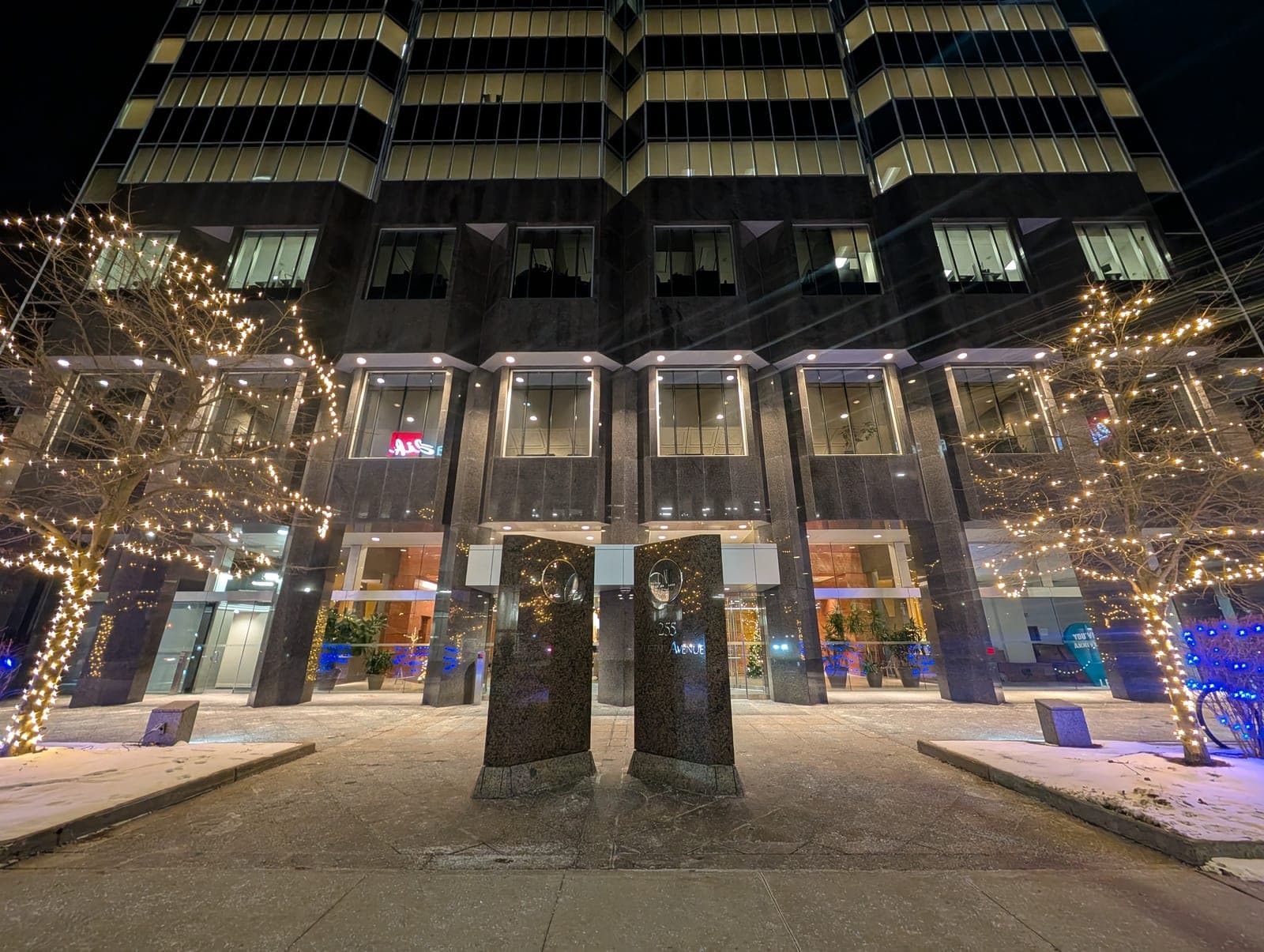Commercial office building facade with entrance trees wrapped in warm-white lights and blue accent uplighting.