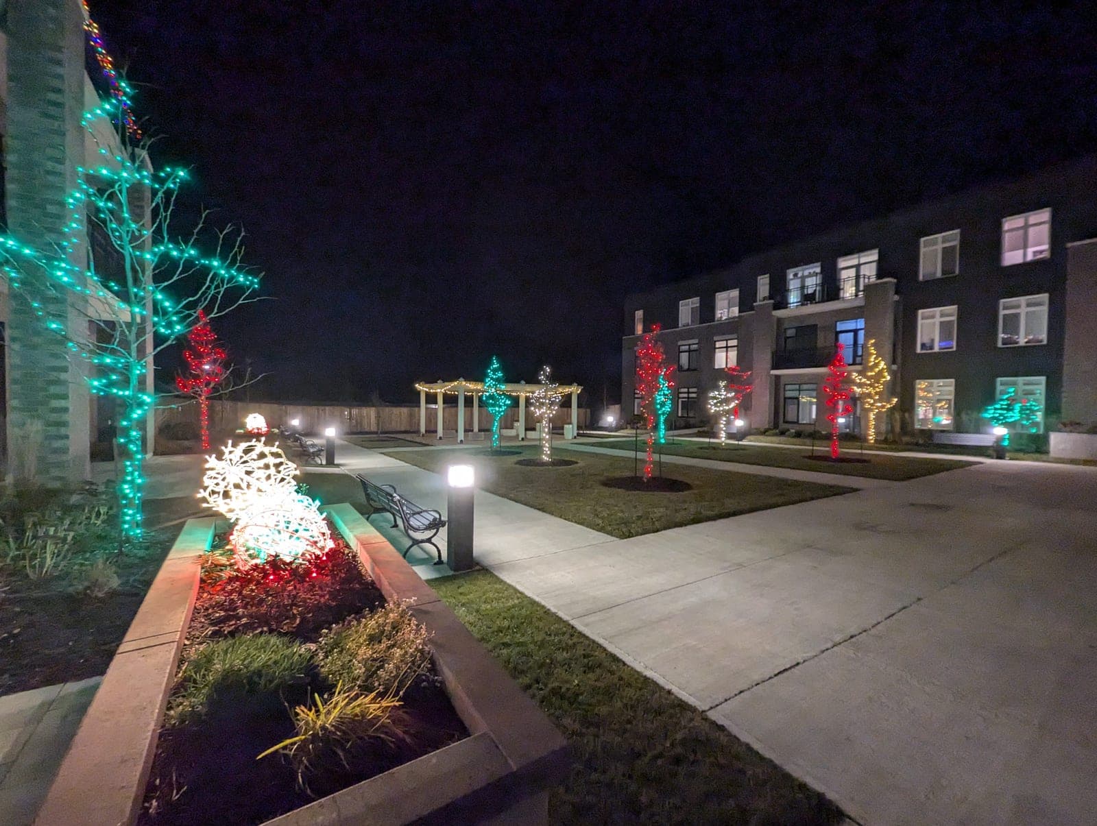 Commercial courtyard walkway with red and green light-wrapped trees and a lit holiday ornament at night