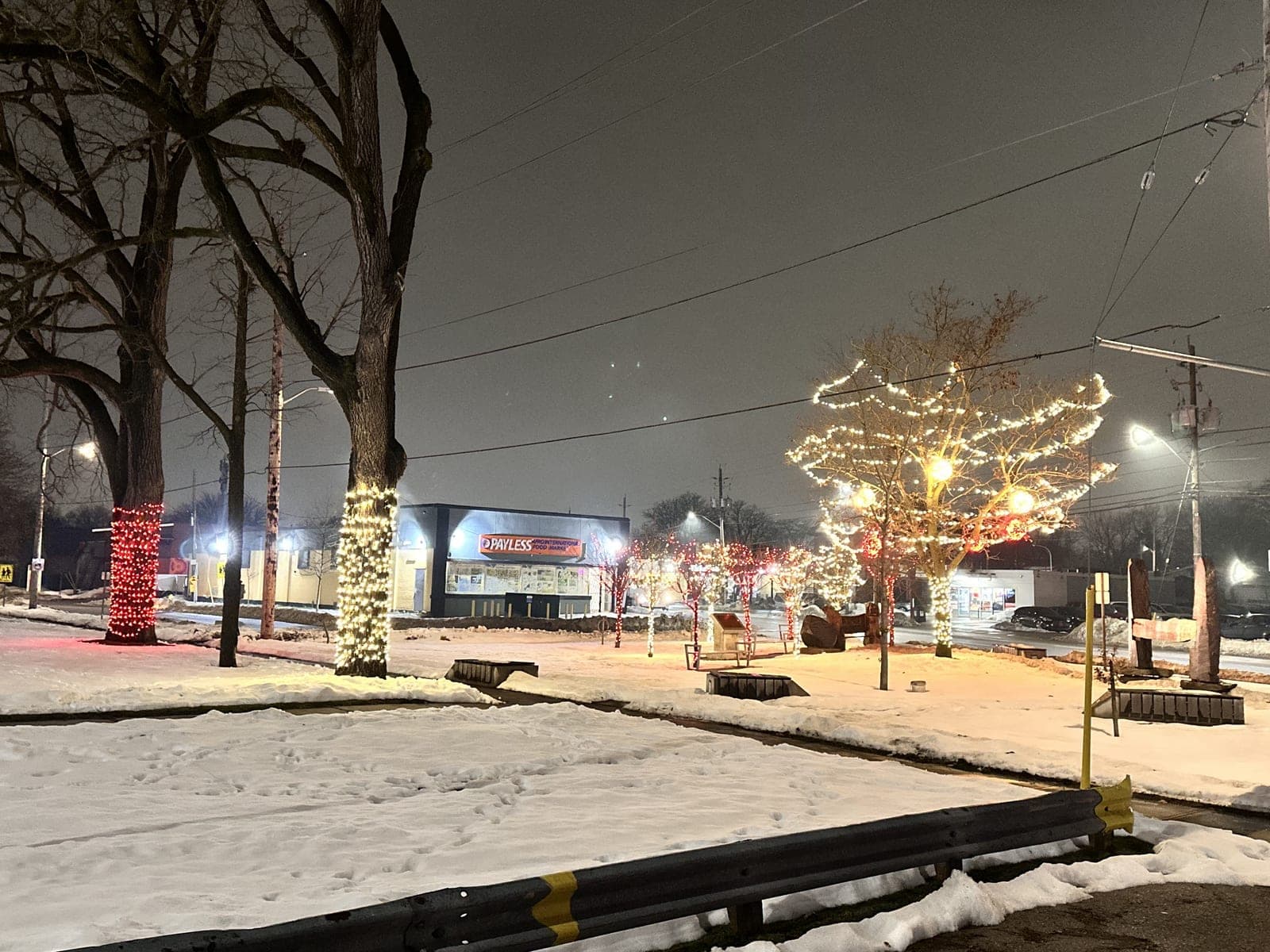Downtown commercial street with bare trees wrapped in warm-white Christmas lights over fresh snow at night