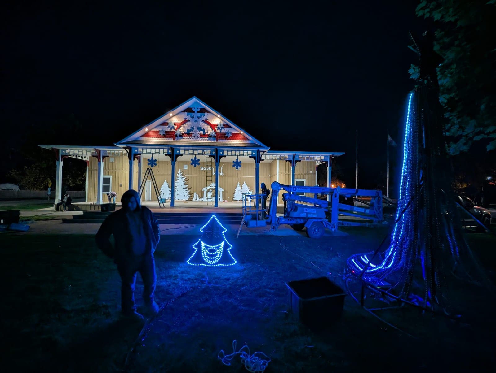 Nighttime holiday light installation at a park pavilion with a boom lift and LED displays.