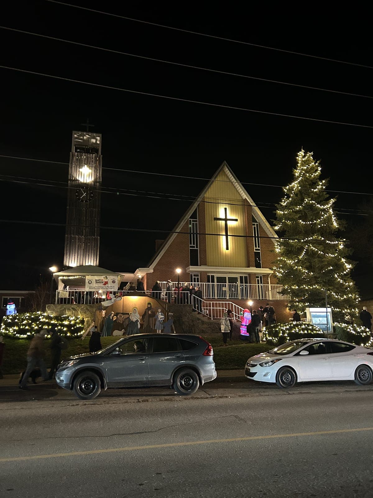 Tall outdoor evergreen wrapped in warm white Christmas lights beside a lit church during a community tree-lighting event at night.