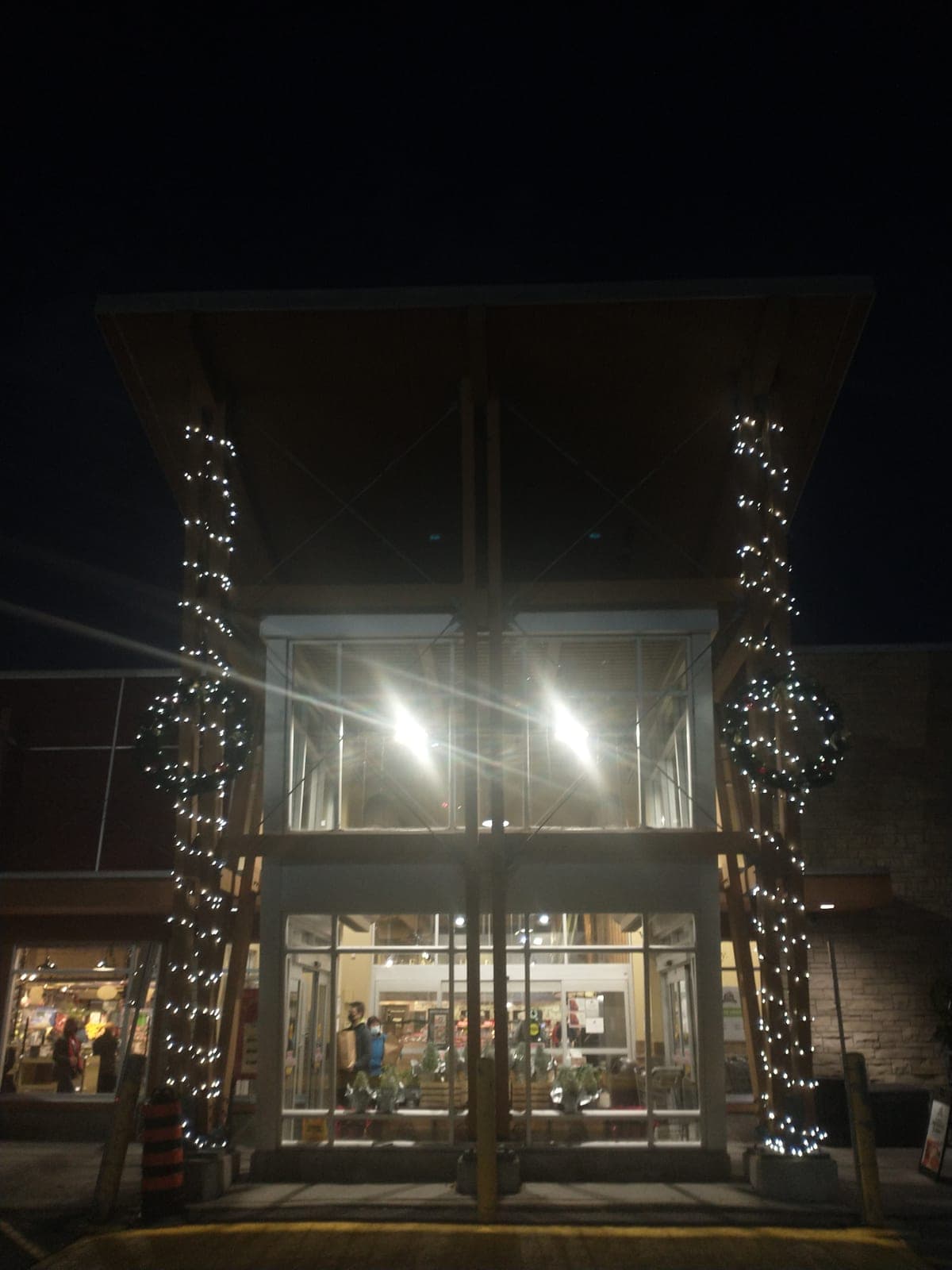 Retail store entrance with tall columns wrapped in Christmas string lights and illuminated wreaths at night.