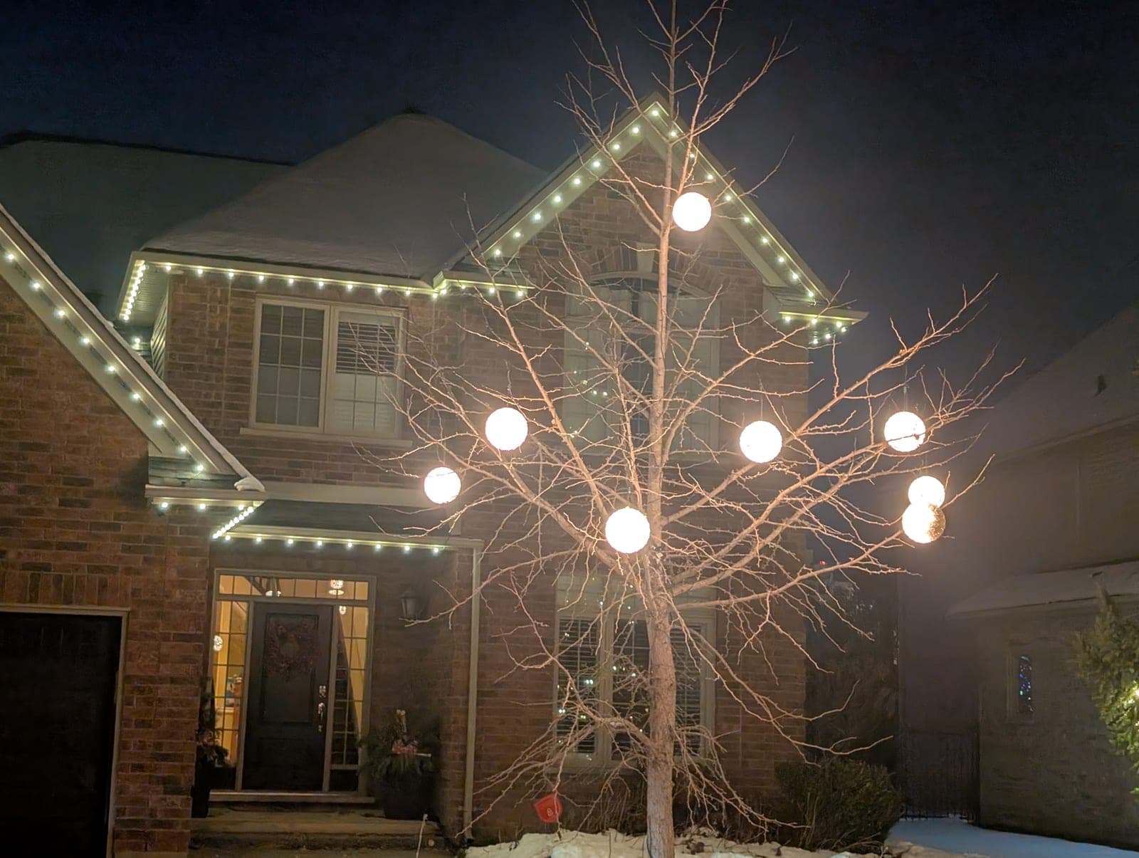 Bare front-yard tree strung with warm-white globe lights beside a brick home with roofline Christmas lighting