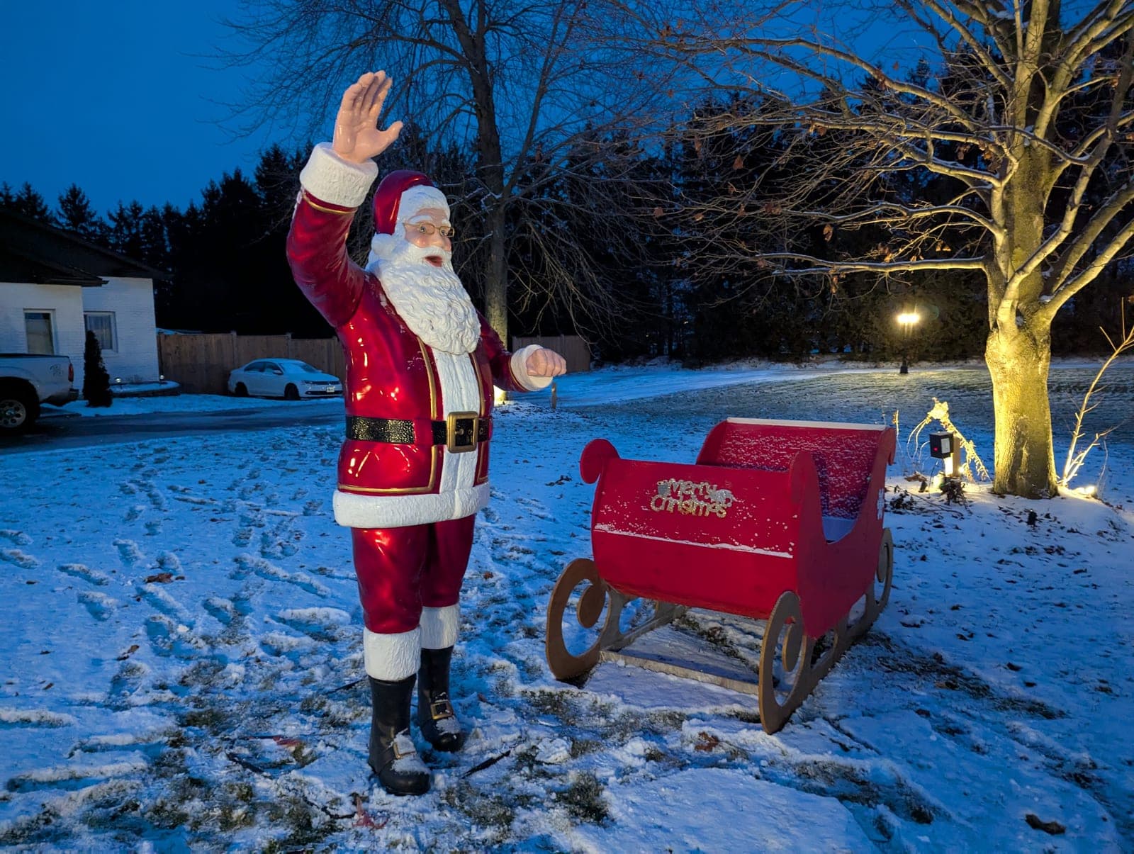Waving Santa figure beside a red sleigh on a snowy yard with a warm-white lit tree at dusk